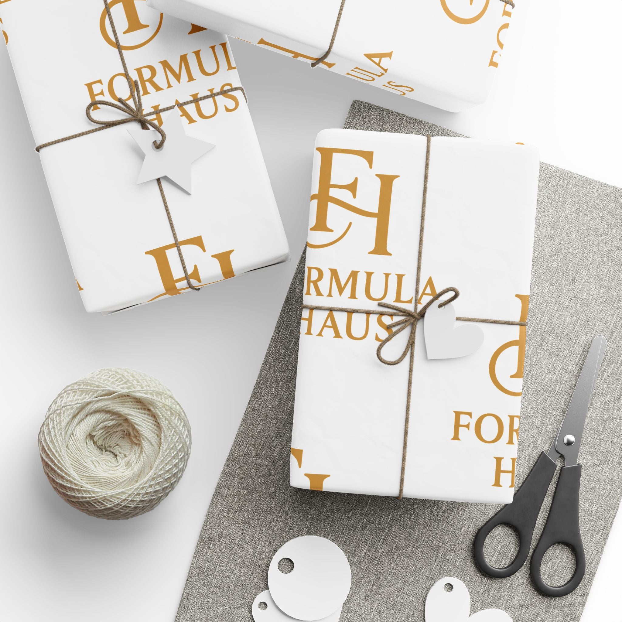 Gift boxes wrapped in white Formula Haus paper with gold monogram logos, tied with twine and star tags, displayed with a spool of twine, blank tags, and scissors on a light grey surface.