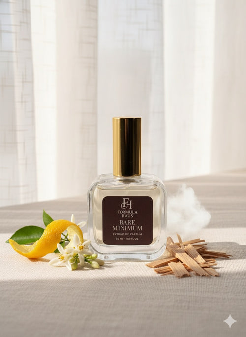 Formula Haus Bare Minimum perfume bottle with a gold cap on a linen surface next to a twisted lemon peel, white flowers, and small wood chips, with sunlight streaming through curtains in the background