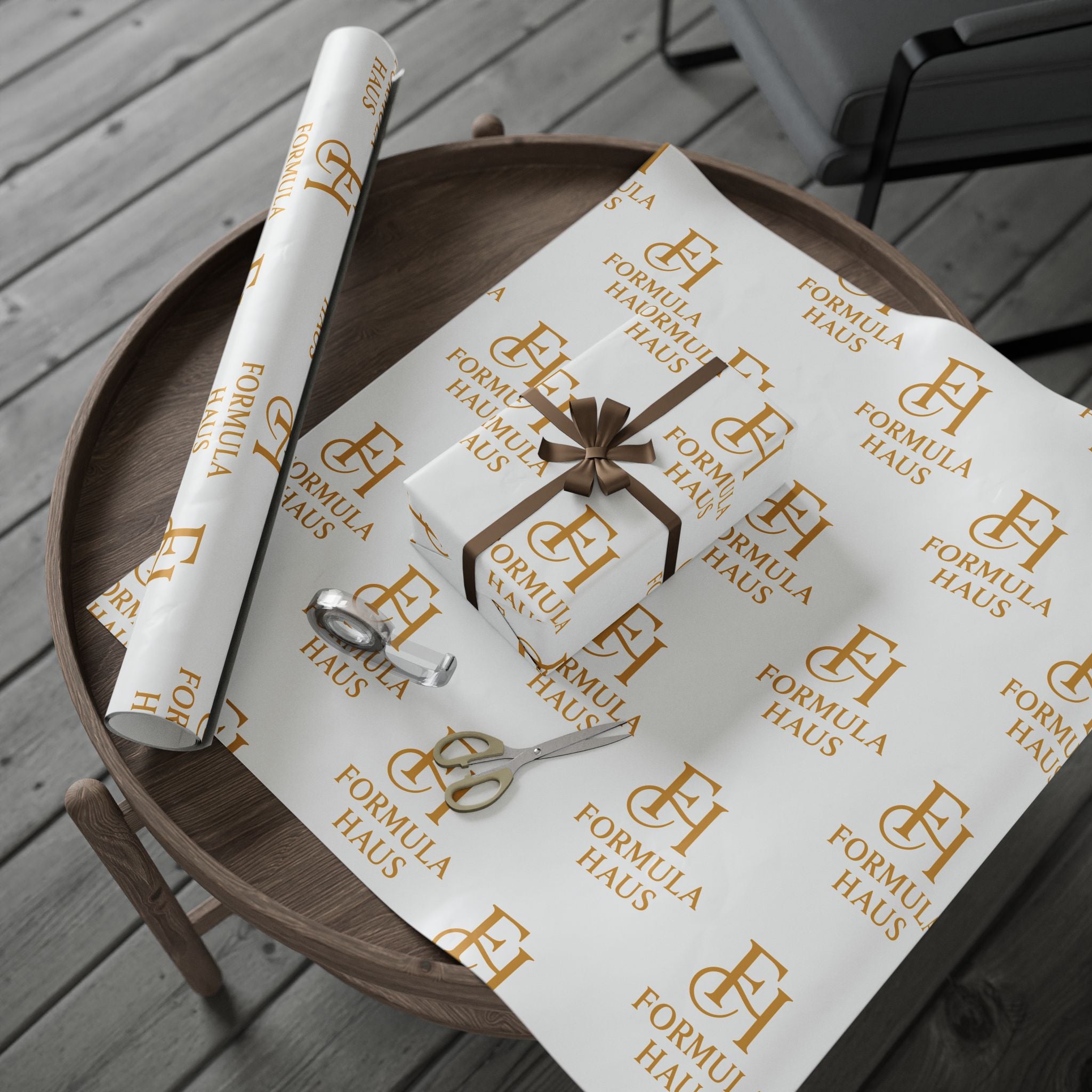 Round wooden tray holding a sheet of white Formula Haus wrapping paper with gold monogram logos, a small gift box wrapped in the same paper with a golden bow, a roll of wrapping paper, and a pair of gold scissors, placed on a wooden deck.