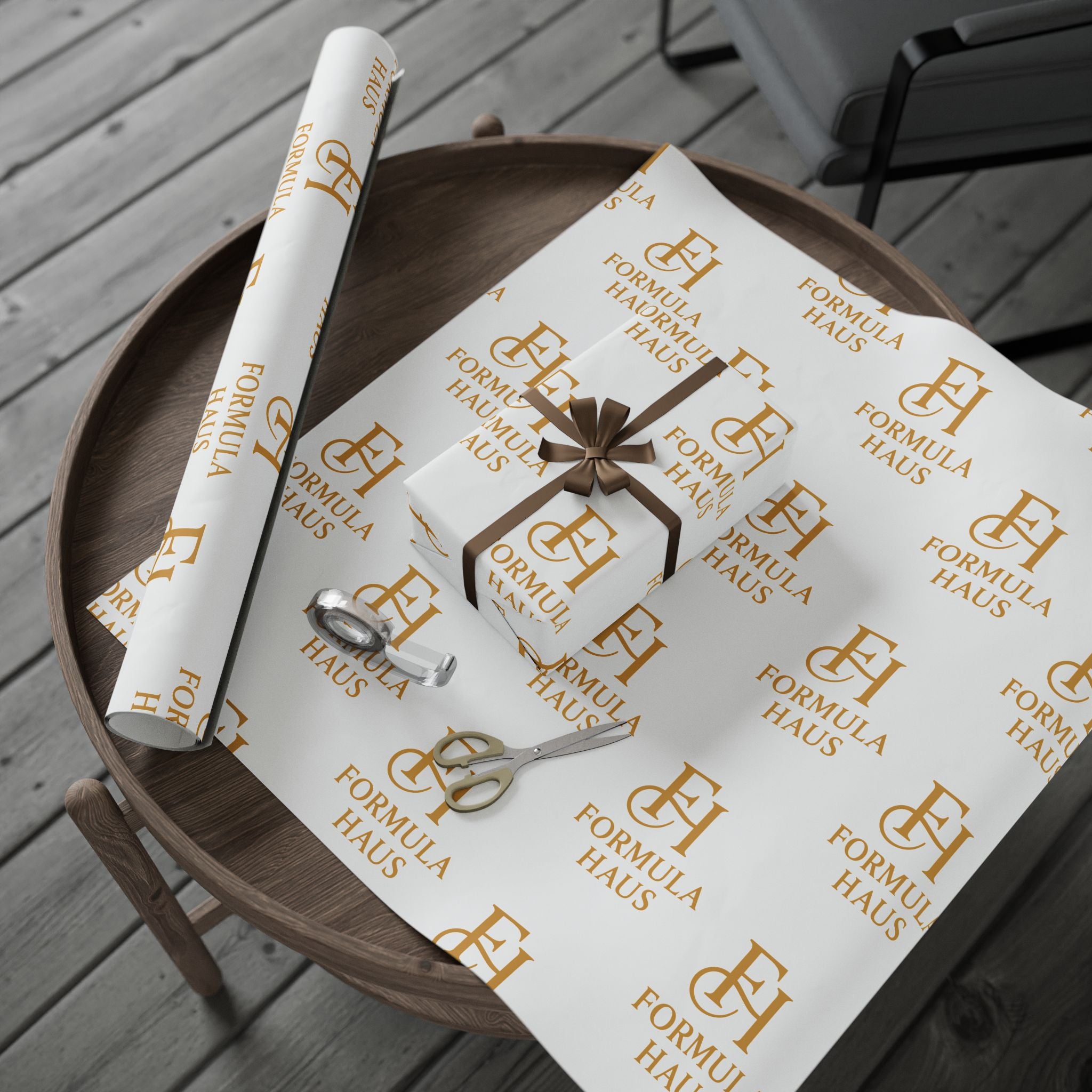 Round wooden tray holding a sheet of white Formula Haus wrapping paper with gold monogram logos, a small gift box wrapped in the same paper with a gold bow, a roll of wrapping paper, and a pair of gold scissors on a wooden deck.