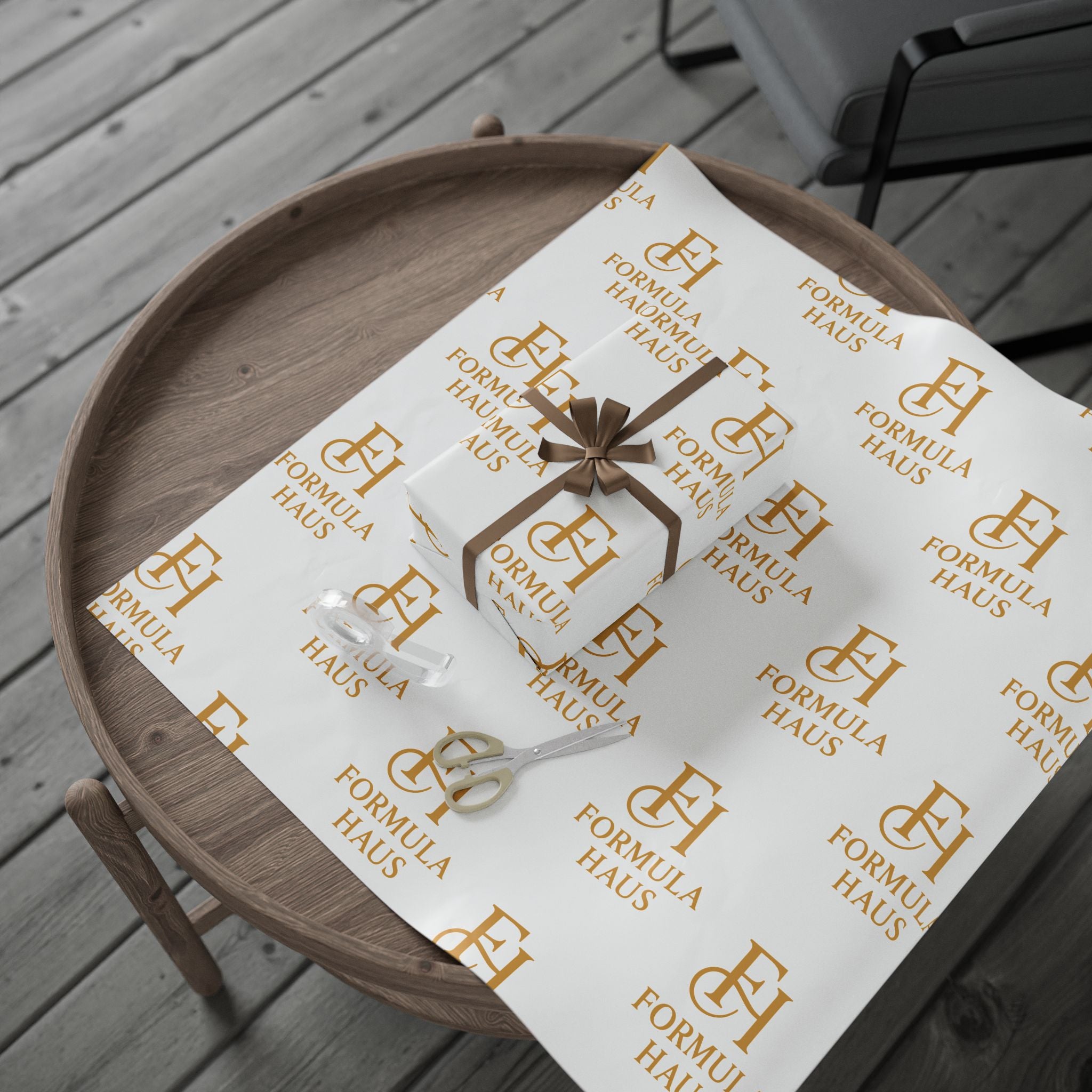 Wooden tray with a small gift box wrapped in white Formula Haus wrapping paper with gold monogram logos, placed on a matching sheet with a pair of silver scissors and ribbon on a wooden floor.